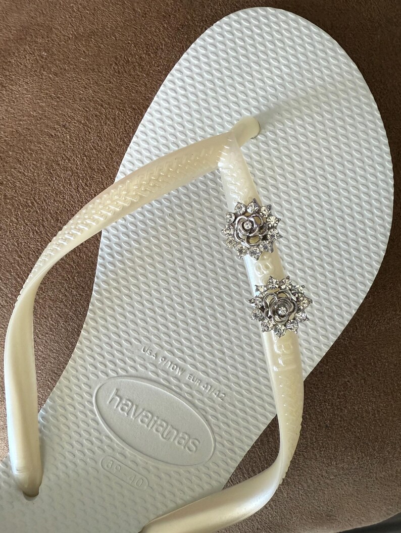 以下が含まれることがあります： 白のハワイアナスのビーチサンダル。ソールはテクスチャード加工で、クリーム色のストラップが付いています。ストラップには2つの銀色の花の装飾が施されています。サイズはEUR 41/42です。