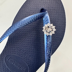 Pode incluir: Um par de chinelos azuis com um charme de flor prateada. Os chinelos t&ecirc;m a palavra "havaianas" impressa na tira e o nome da marca "havaianas" na sola. A sola tamb&eacute;m tem o tamanho "37/38" impresso.