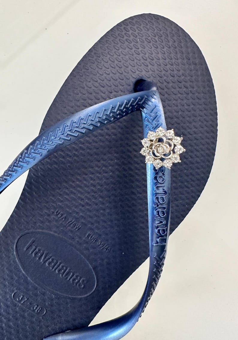 Pode incluir: Um par de chinelos azuis com um charme de flor prateada. Os chinelos t&ecirc;m a palavra "havaianas" impressa na tira e o nome da marca "havaianas" na sola. A sola tamb&eacute;m tem o tamanho "37/38" impresso.