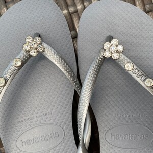 May include: A pair of gray flip-flops with a silver finish. The flip-flops have a flower-shaped rhinestone embellishment on each strap. The straps are labeled "Seut Nous" and the flip-flops have the brand name "havaianas" printed on the soles.