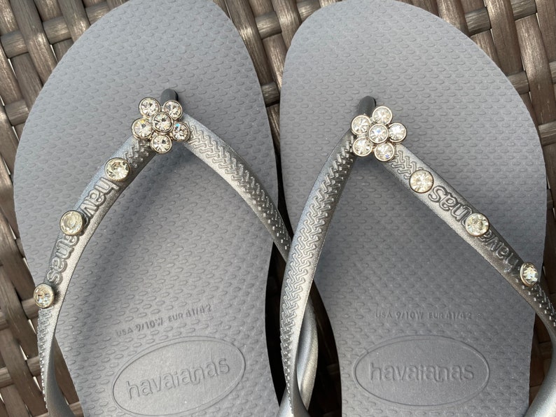 May include: A pair of gray flip-flops with a silver finish. The flip-flops have a flower-shaped rhinestone embellishment on each strap. The straps are labeled "Seut Nous" and the flip-flops have the brand name "havaianas" printed on the soles.