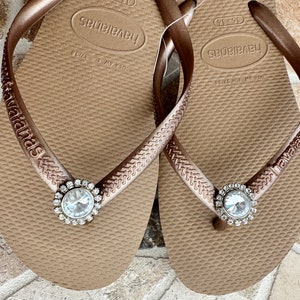May include: A pair of brown Havaianas flip-flops with a sparkling, round, clear rhinestone embellishment. The straps are a matching brown colour with the brand name embossed. The flip-flops are a size EUR 37/38.