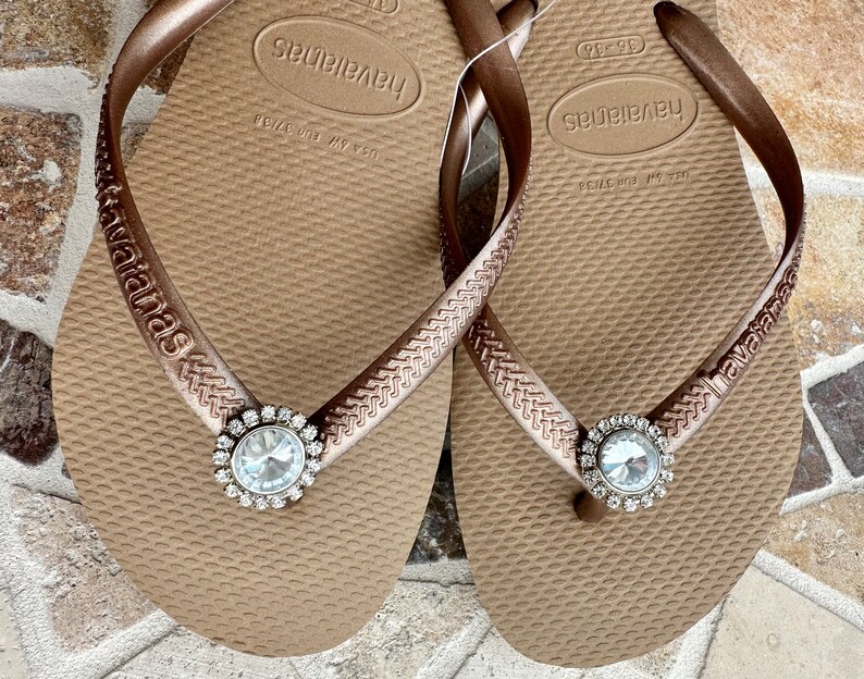 May include: A pair of brown Havaianas flip-flops with a sparkling, round, clear rhinestone embellishment. The straps are a matching brown colour with the brand name embossed. The flip-flops are a size EUR 37/38.
