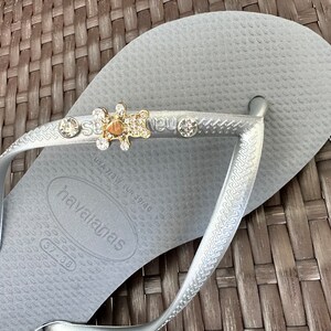 Puede incluir: Chanclas plateadas con un dije de oso brillante y pedrer&iacute;a. Las chanclas tienen el nombre de la marca "havaianas" impreso en la suela.