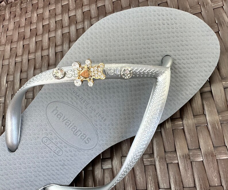Puede incluir: Chanclas plateadas con un dije de oso brillante y pedrer&iacute;a. Las chanclas tienen el nombre de la marca "havaianas" impreso en la suela.