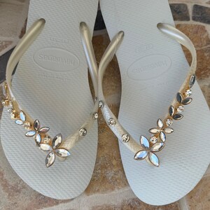 May include: A pair of white Havaianas flip-flops with clear straps. The straps are adorned with a floral design of gold, silver, and clear gemstones. The flip-flops are displayed on a patterned surface.