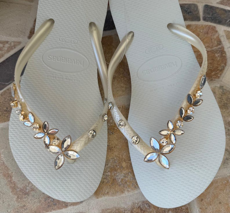 May include: A pair of white Havaianas flip-flops with clear straps. The straps are adorned with a floral design of gold, silver, and clear gemstones. The flip-flops are displayed on a patterned surface.