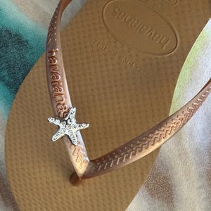 K&ouml;nnte beinhalten: Hellbraune Havaianas-Zehensandalen mit einem silbernen Seestern-Anh&auml;nger am Riemen. Die Sohle ist strukturiert und tr&auml;gt den Markennamen. Diese l&auml;ssigen Flip-Flops sind ideal f&uuml;r den Sommer.