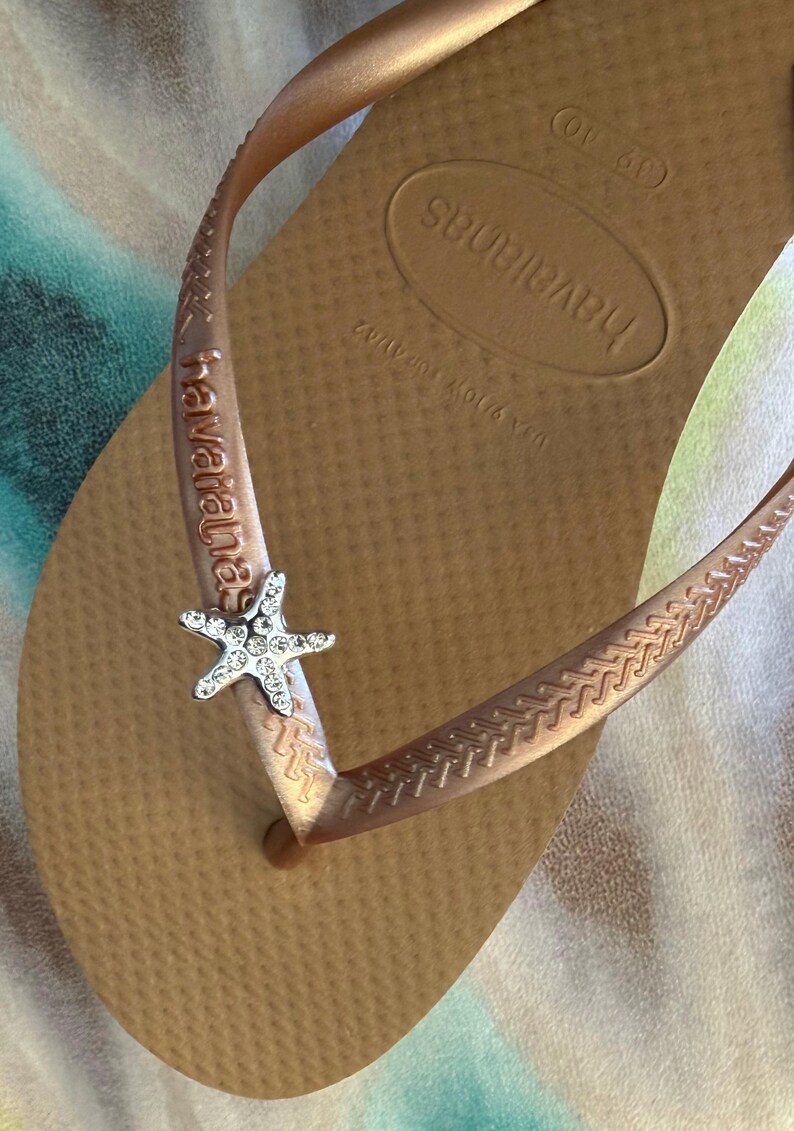 K&ouml;nnte beinhalten: Hellbraune Havaianas-Zehensandalen mit einem silbernen Seestern-Anh&auml;nger am Riemen. Die Sohle ist strukturiert und tr&auml;gt den Markennamen. Diese l&auml;ssigen Flip-Flops sind ideal f&uuml;r den Sommer.