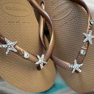 K&ouml;nnte beinhalten: Bronze farbene Havaianas-Zehensandalen mit verzierten Riemen. Die Riemen sind mit silbernen Seesternen und klaren Kristallakzenten verziert. Gr&ouml;&szlig;e EUR 37/38.