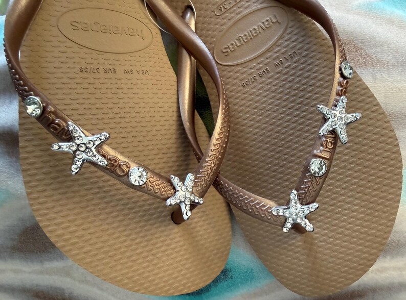 K&ouml;nnte beinhalten: Bronze farbene Havaianas-Zehensandalen mit verzierten Riemen. Die Riemen sind mit silbernen Seesternen und klaren Kristallakzenten verziert. Gr&ouml;&szlig;e EUR 37/38.