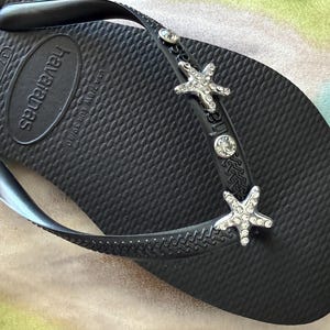 K&ouml;nnte beinhalten: Schwarze Havaianas-Zehensandale mit strukturierter Oberfl&auml;che. Der Riemen ist mit silbernen Seesternen und klaren Kristallakzenten verziert. Der Markenname ist auf dem Fu&szlig;bett gepr&auml;gt.