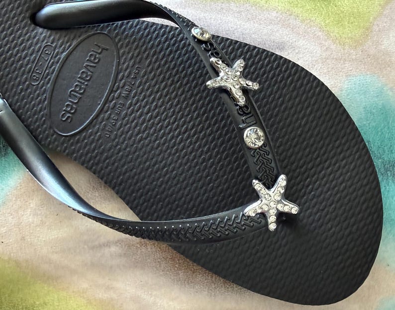 K&ouml;nnte beinhalten: Schwarze Havaianas-Zehensandale mit strukturierter Oberfl&auml;che. Der Riemen ist mit silbernen Seesternen und klaren Kristallakzenten verziert. Der Markenname ist auf dem Fu&szlig;bett gepr&auml;gt.