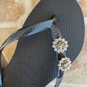 以下が含まれることがあります： 黒いハワイアナスのビーチサンダル、銀色の花の装飾付き。ビーチサンダルは、テクスチャード加工されたフットベッドと黒いストラップが付いています。装飾は、花の形に配置された小さな輝くクリスタルで作られています。