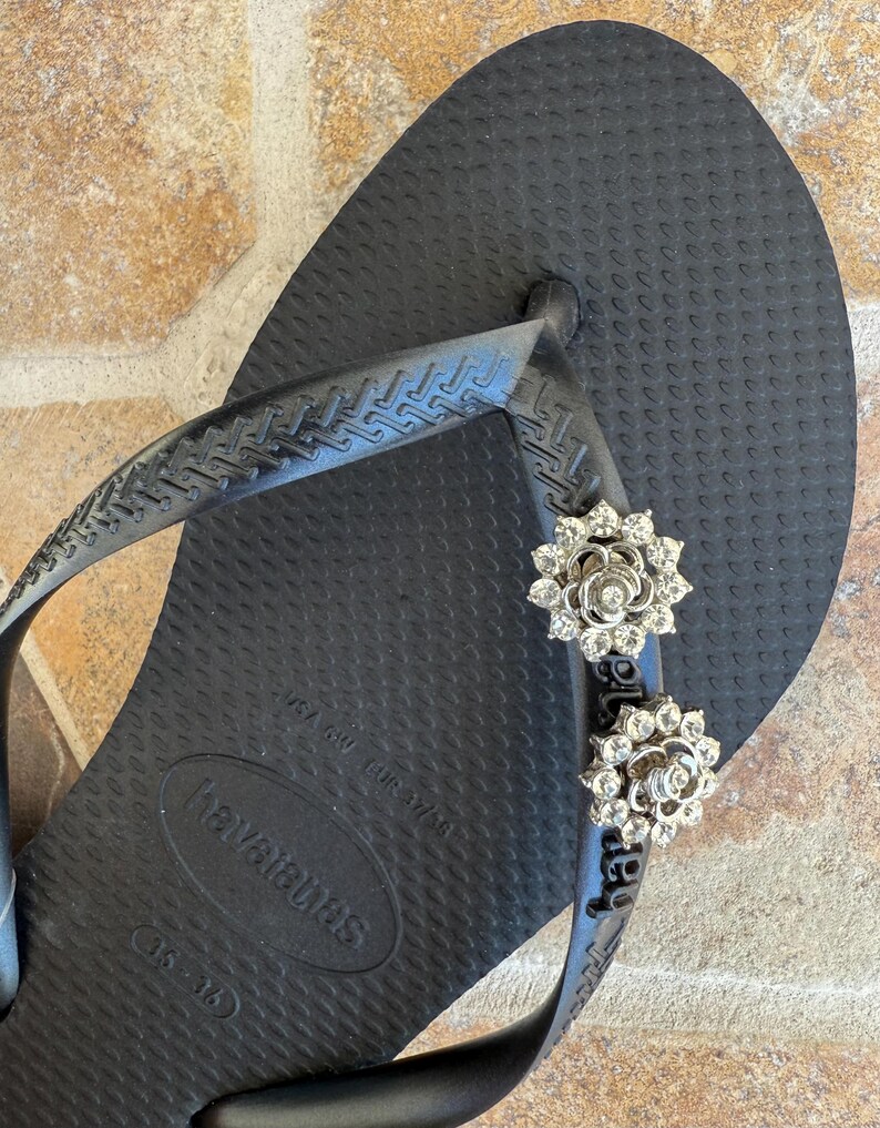 以下が含まれることがあります： 黒いハワイアナスのビーチサンダル、銀色の花の装飾付き。ビーチサンダルは、テクスチャード加工されたフットベッドと黒いストラップが付いています。装飾は、花の形に配置された小さな輝くクリスタルで作られています。