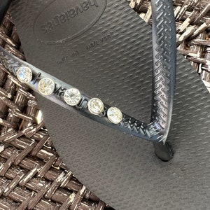 May include: Black flip-flop sandals with a textured sole and a single strap adorned with rhinestones. The sandals have the brand name "Havaianas" printed on the sole. The size is marked as "USA 7/8W EUR 39/40".