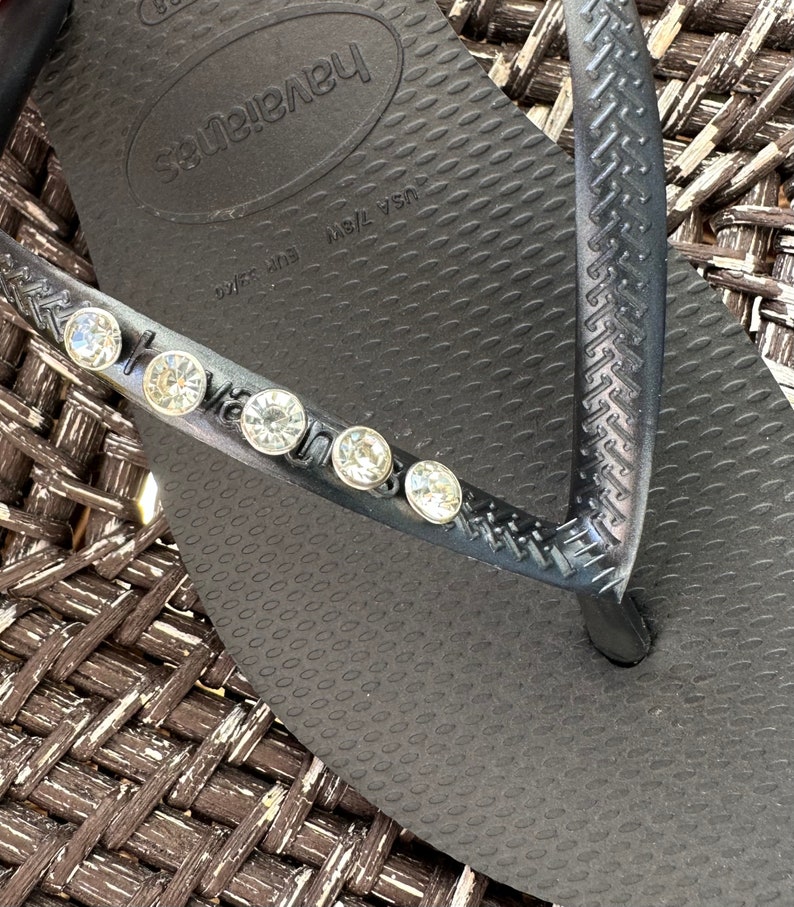 May include: Black flip-flop sandals with a textured sole and a single strap adorned with rhinestones. The sandals have the brand name "Havaianas" printed on the sole. The size is marked as "USA 7/8W EUR 39/40".
