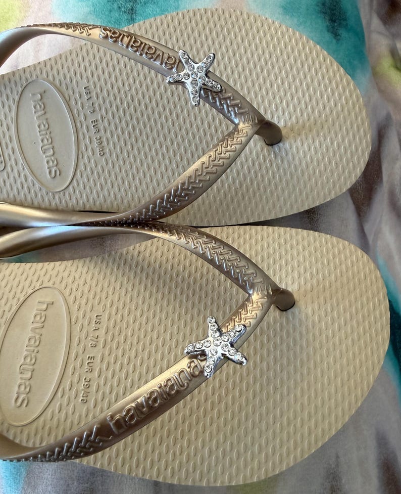 K&ouml;nnte beinhalten: Ein Paar beige Havaianas-Flip-Flops mit silbernen Riemen und einem Strass-Seestern-Besatz. Die Flip-Flops sind in der Gr&ouml;&szlig;e EUR 39.