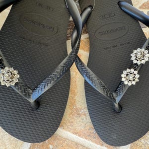 以下が含まれることがあります： 黒いハワイアナスのビーチサンダル、銀色の花の装飾付き。ビーチサンダルは、テクスチャード加工されたフットベッドと、クラシックなトングストラップのデザインが特徴です。装飾は銀色の金属と透明なラインストーンで作られています。