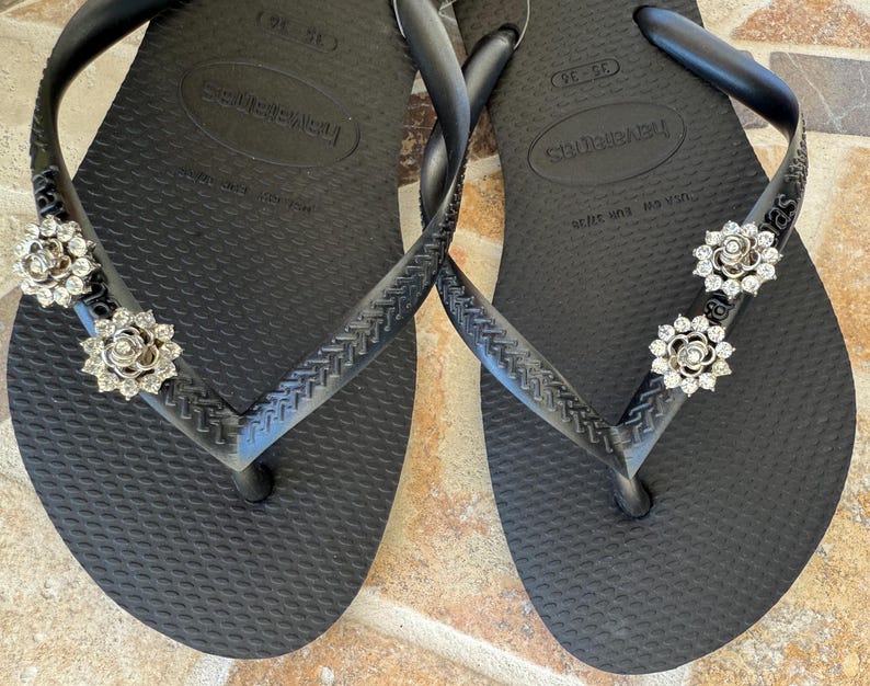 以下が含まれることがあります： 黒いハワイアナスのビーチサンダル、銀色の花の装飾付き。ビーチサンダルは、テクスチャード加工されたフットベッドと、クラシックなトングストラップのデザインが特徴です。装飾は銀色の金属と透明なラインストーンで作られています。