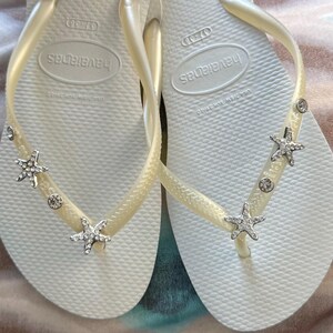 K&ouml;nnte beinhalten: Ein Paar wei&szlig;e Havaianas-Flip-Flops mit cremefarbenen Riemen. Jeder Riemen ist mit silbernen Seesternen und runden, klaren Edelsteinen verziert. Die Flip-Flops sind in der Gr&ouml;&szlig;e 37-38.