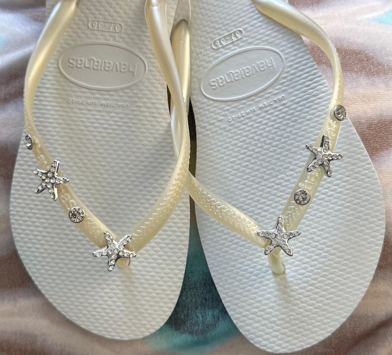 K&ouml;nnte beinhalten: Ein Paar wei&szlig;e Havaianas-Flip-Flops mit cremefarbenen Riemen. Jeder Riemen ist mit silbernen Seesternen und runden, klaren Edelsteinen verziert. Die Flip-Flops sind in der Gr&ouml;&szlig;e 37-38.