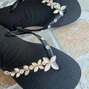 May include: Black flip-flops with a floral design. The straps are adorned with clear rhinestones and a gold chain. The flowers are iridescent with clear crystal centers. The flip-flops are on a textured surface.