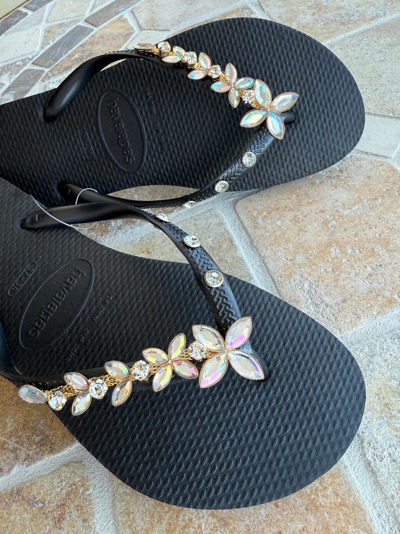 May include: Black flip-flops with a floral design. The straps are adorned with clear rhinestones and a gold chain. The flowers are iridescent with clear crystal centers. The flip-flops are on a textured surface.