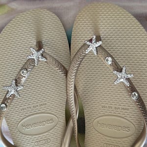 K&ouml;nnte beinhalten: Ein Paar beige Havaianas-Flip-Flops mit silbernen Seesternen und Strasssteinen auf den Riemen. Die Flip-Flops sind mit den Gr&ouml;&szlig;en USA 7/8 und EUR 39/40 gekennzeichnet.