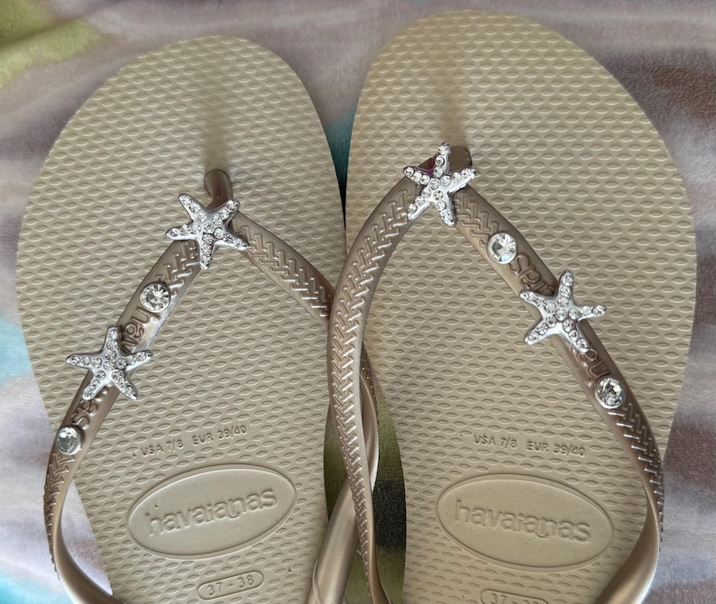 K&ouml;nnte beinhalten: Ein Paar beige Havaianas-Flip-Flops mit silbernen Seesternen und Strasssteinen auf den Riemen. Die Flip-Flops sind mit den Gr&ouml;&szlig;en USA 7/8 und EUR 39/40 gekennzeichnet.