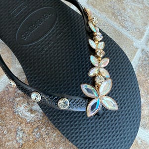 May include: Black flip-flop with a decorative floral design. The flip-flop features a gold-colored metal accent with iridescent crystal-like flower embellishments. Additional clear crystals are set along the strap.