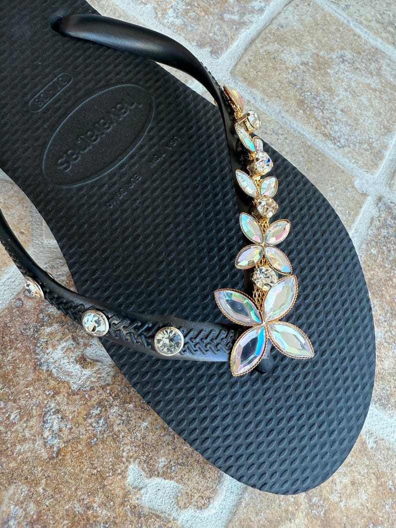 May include: Black flip-flop with a decorative floral design. The flip-flop features a gold-colored metal accent with iridescent crystal-like flower embellishments. Additional clear crystals are set along the strap.