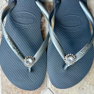May include: Dark grey Havaianas flip-flops with silver straps. Each flip-flop has a large, clear rhinestone embellishment at the toe post. The Havaianas logo is visible on the strap.