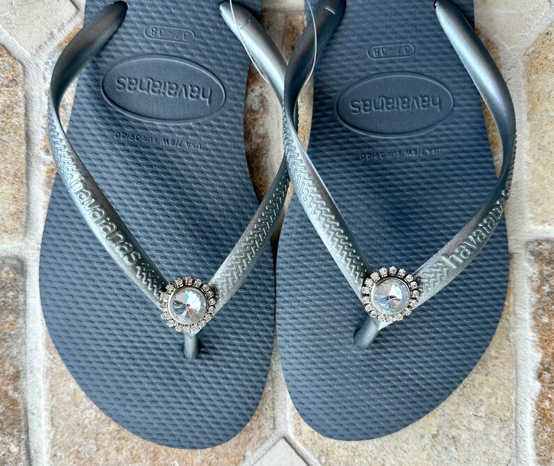 May include: Dark grey Havaianas flip-flops with silver straps. Each flip-flop has a large, clear rhinestone embellishment at the toe post. The Havaianas logo is visible on the strap.