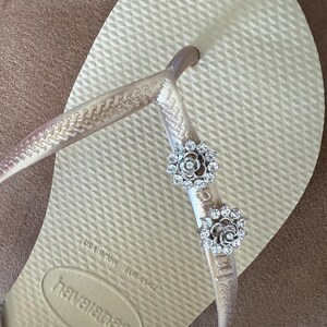 以下が含まれることがあります： ベージュ色のハワイアナスのビーチサンダル。透明なストラップに、2つの銀色の花の装飾が施されています。サンダルはEUR 41/42サイズです。