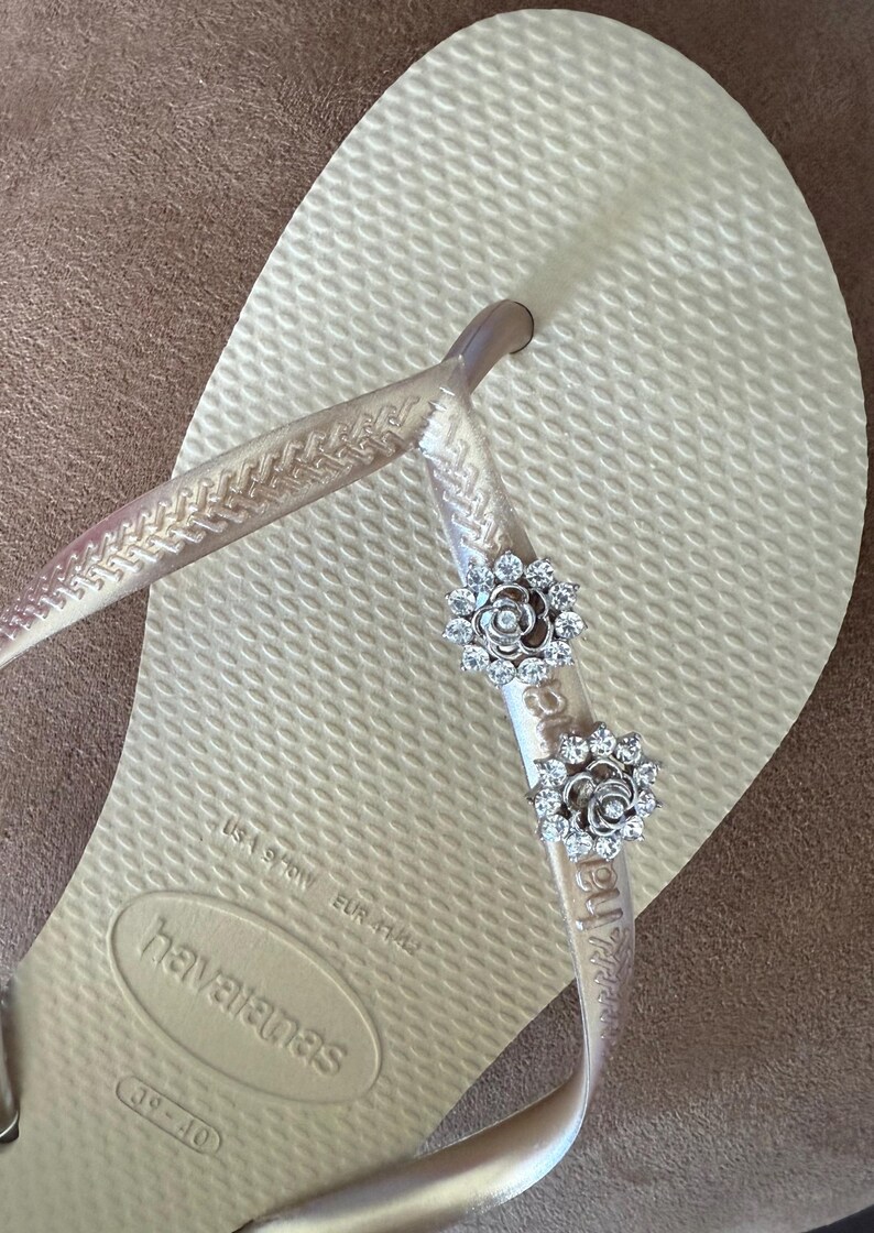 以下が含まれることがあります： ベージュ色のハワイアナスのビーチサンダル。透明なストラップに、2つの銀色の花の装飾が施されています。サンダルはEUR 41/42サイズです。