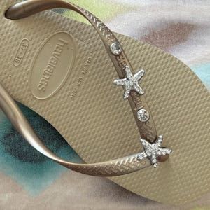 K&ouml;nnte beinhalten: Nahaufnahme einer beigefarbenen Havaianas-Zehensandale mit einem metallischen Riemen, der mit Strass-Seesternen und klaren Strasssteinen verziert ist. Die Zehensandale ist Gr&ouml;&szlig;e 39/40 EUR.