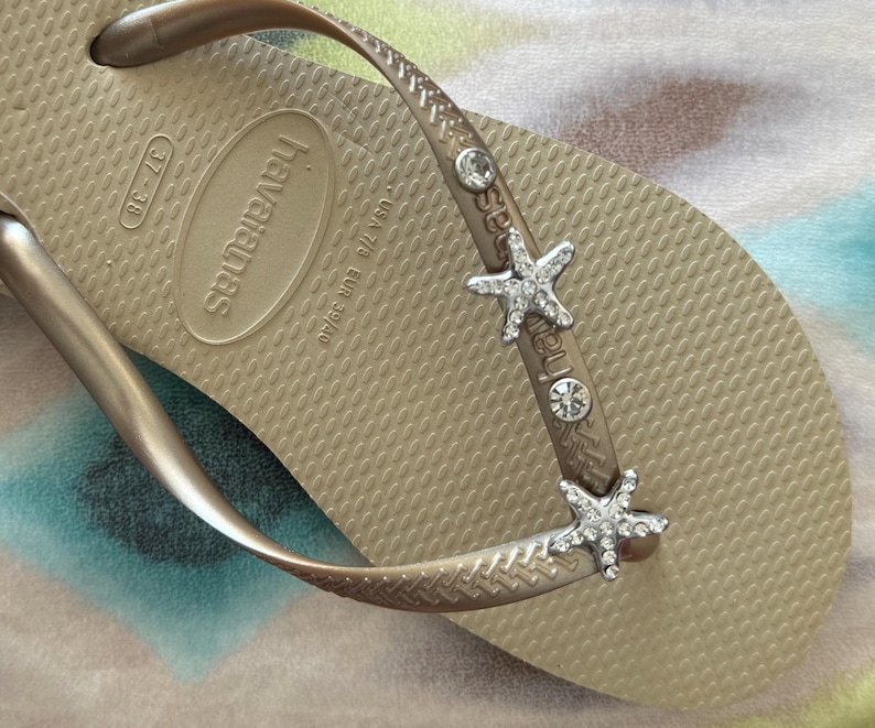 K&ouml;nnte beinhalten: Nahaufnahme einer beigefarbenen Havaianas-Zehensandale mit einem metallischen Riemen, der mit Strass-Seesternen und klaren Strasssteinen verziert ist. Die Zehensandale ist Gr&ouml;&szlig;e 39/40 EUR.