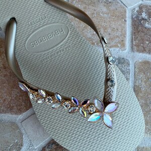 May include: Tan Havaianas flip-flop with a gold strap adorned with iridescent and clear crystal embellishments. The decorative accents are arranged in a floral pattern, adding a touch of glamour to the casual footwear.
