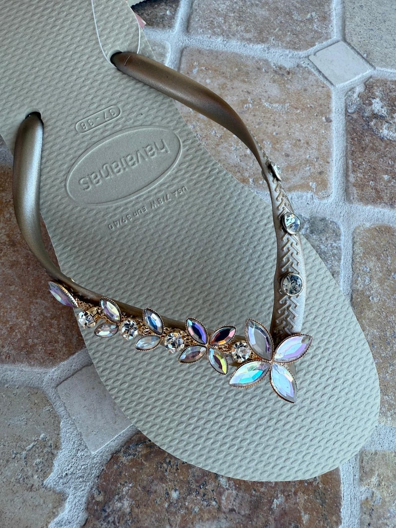May include: Tan Havaianas flip-flop with a gold strap adorned with iridescent and clear crystal embellishments. The decorative accents are arranged in a floral pattern, adding a touch of glamour to the casual footwear.