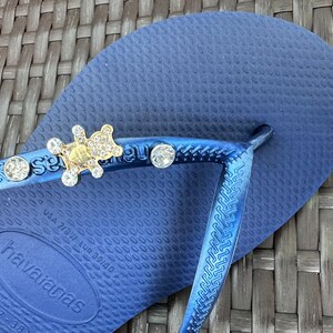 Puede incluir: Chanclas azules con un dije de oso de oro y cristal. Las chanclas tienen el texto "Havaianas" en la suela.
