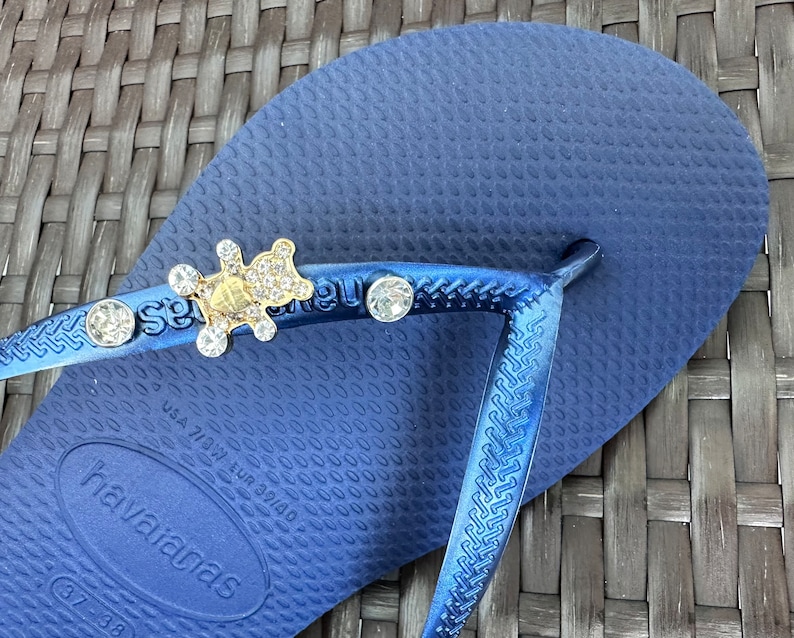 Puede incluir: Chanclas azules con un dije de oso de oro y cristal. Las chanclas tienen el texto "Havaianas" en la suela.