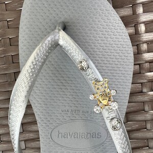 Puede incluir: Chanclas de dedo plateadas con un dije de oso dorado y cristal. Las chanclas tienen el nombre de la marca "havaianas" y la talla "37-38" impresa en la suela.