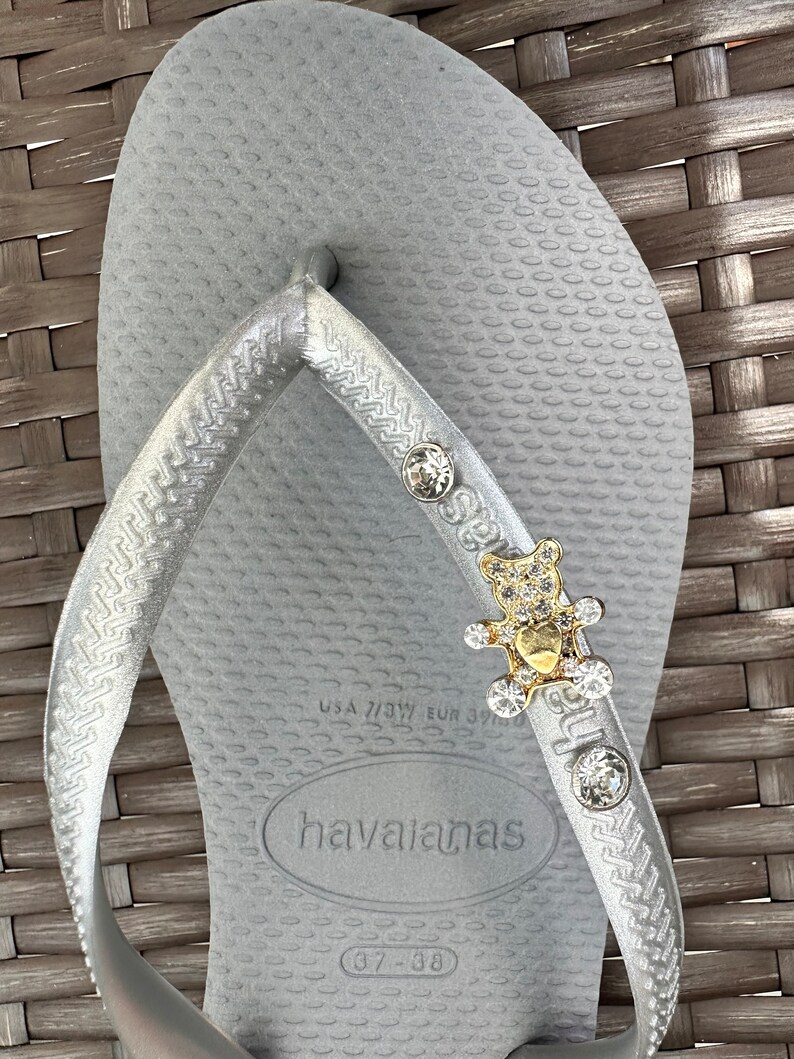 Puede incluir: Chanclas de dedo plateadas con un dije de oso dorado y cristal. Las chanclas tienen el nombre de la marca "havaianas" y la talla "37-38" impresa en la suela.