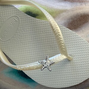 K&ouml;nnte beinhalten: Wei&szlig;e Havaianas-Zehensandale mit einem silbernen Seestern-Charm. Die Zehensandale hat eine strukturierte Oberfl&auml;che und einen perlmuttfarbenen Riemen. Das Havaianas-Logo ist auf dem Fu&szlig;bett sichtbar. Ein Accessoire zum Thema Strand.