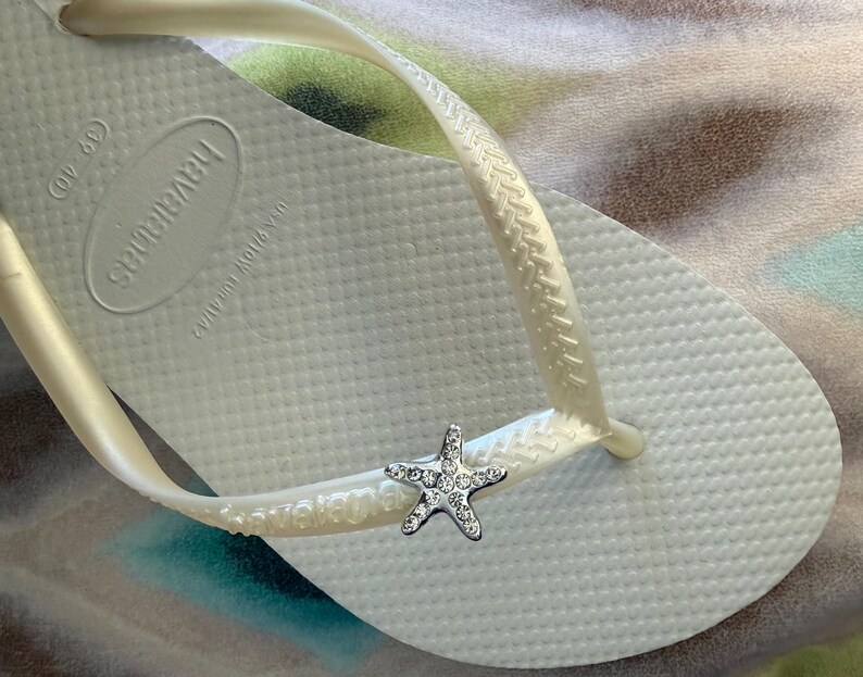 K&ouml;nnte beinhalten: Wei&szlig;e Havaianas-Zehensandale mit einem silbernen Seestern-Charm. Die Zehensandale hat eine strukturierte Oberfl&auml;che und einen perlmuttfarbenen Riemen. Das Havaianas-Logo ist auf dem Fu&szlig;bett sichtbar. Ein Accessoire zum Thema Strand.