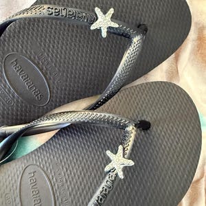 K&ouml;nnte beinhalten: Dunkelgraue Havaianas-Flip-Flops mit strukturierter Oberfl&auml;che. Jeder Riemen hat einen silberfarbenen Seestern-Anh&auml;nger. Das Havaianas-Logo ist auf Fu&szlig;bett und Riemen sichtbar. Gr&ouml;&szlig;e 39-40.