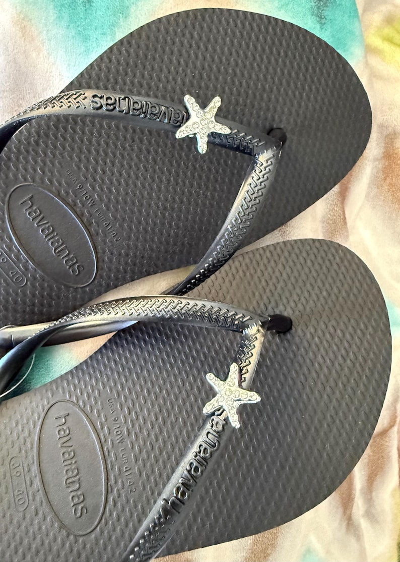 K&ouml;nnte beinhalten: Dunkelgraue Havaianas-Flip-Flops mit strukturierter Oberfl&auml;che. Jeder Riemen hat einen silberfarbenen Seestern-Anh&auml;nger. Das Havaianas-Logo ist auf Fu&szlig;bett und Riemen sichtbar. Gr&ouml;&szlig;e 39-40.