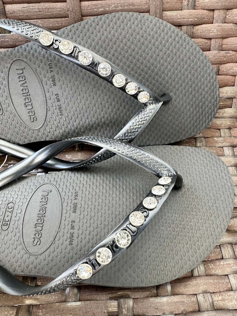 May include: Black flip-flops with a rhinestone embellishment on the strap. The flip-flops have the text "havaianas" on the sole. The size is marked as "USA 7/8W EUR 39/40".