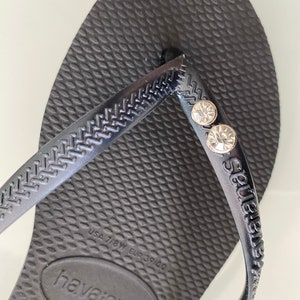 May include: Black Havaianas flip-flops with a textured sole and straps. The straps feature two clear, round embellishments. The brand name is embossed on the strap and sole. The size is USA 7/8W, EUR 39/40.